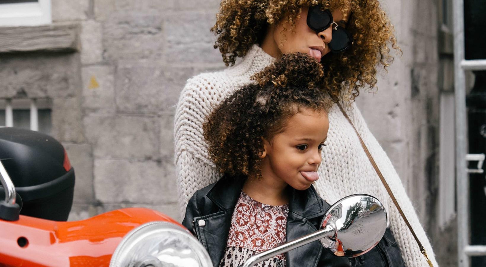 Close to the center of everything mother and daughter stick their tongues out while looking at themselves in a motorcycle mirror parked in front of a building